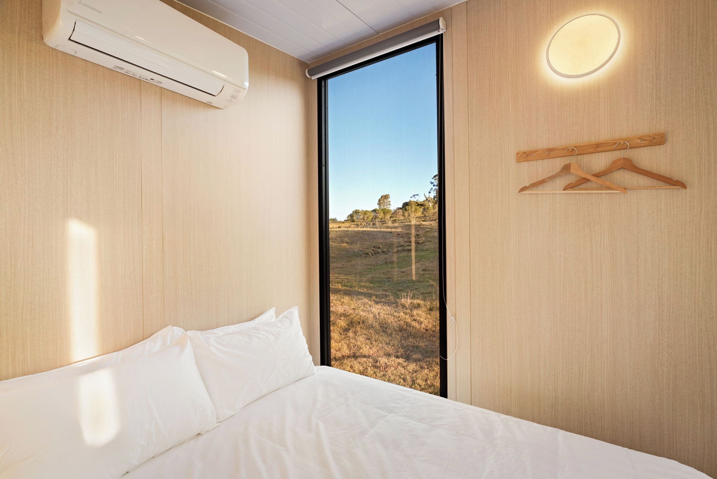 Une chambre à coucher comprenant un lit, un appareil de climatisation, une fenêtre donnant sur un champ et un luminaire fixé au mur.