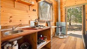 Une petite cabane en bois comprenant un coin cuisine équipé d’un évier, d’une cuisinière et d’étagères.