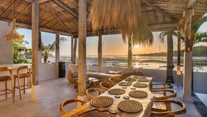 Un restaurant situé en bord de mer, avec une longue table à manger, des chaises en osier et une vue sur l’océan.