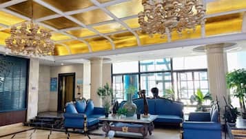 Un vestíbulo de hotel con techo alto, sofás azules, una lámpara de araña grande y ventanas amplias.