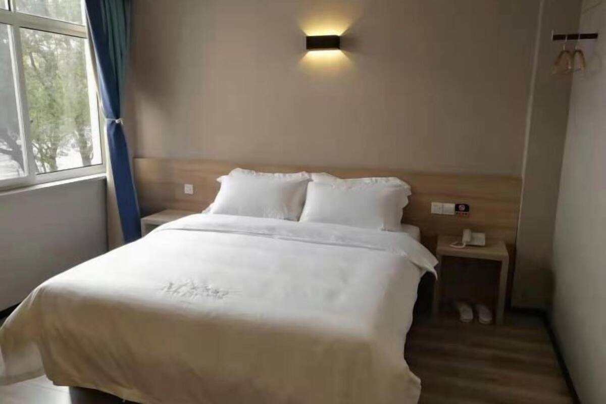 Et hotelværelse med en seng, et natbord, en vægmonteret lampe og et vindue med gardiner.