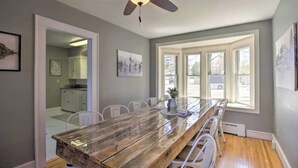 Un comedor con mesa de madera, sillas blancas, ventilador de techo y ventanas amplias.