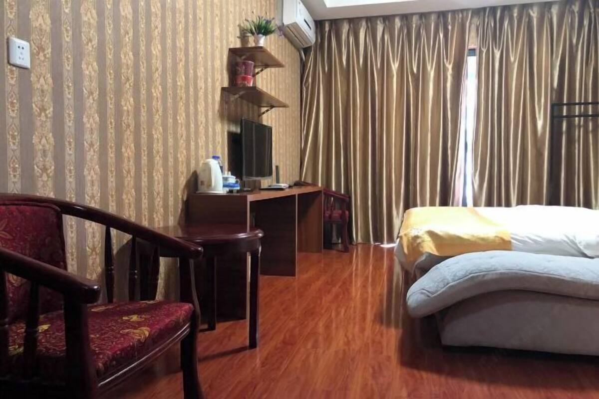 Une chambre d’hôtel avec un plancher en bois, un bureau avec un ordinateur et une chaise, un lit recouvert d’une courtepointe blanche et jaune, et une fenêtre avec des rideaux.
