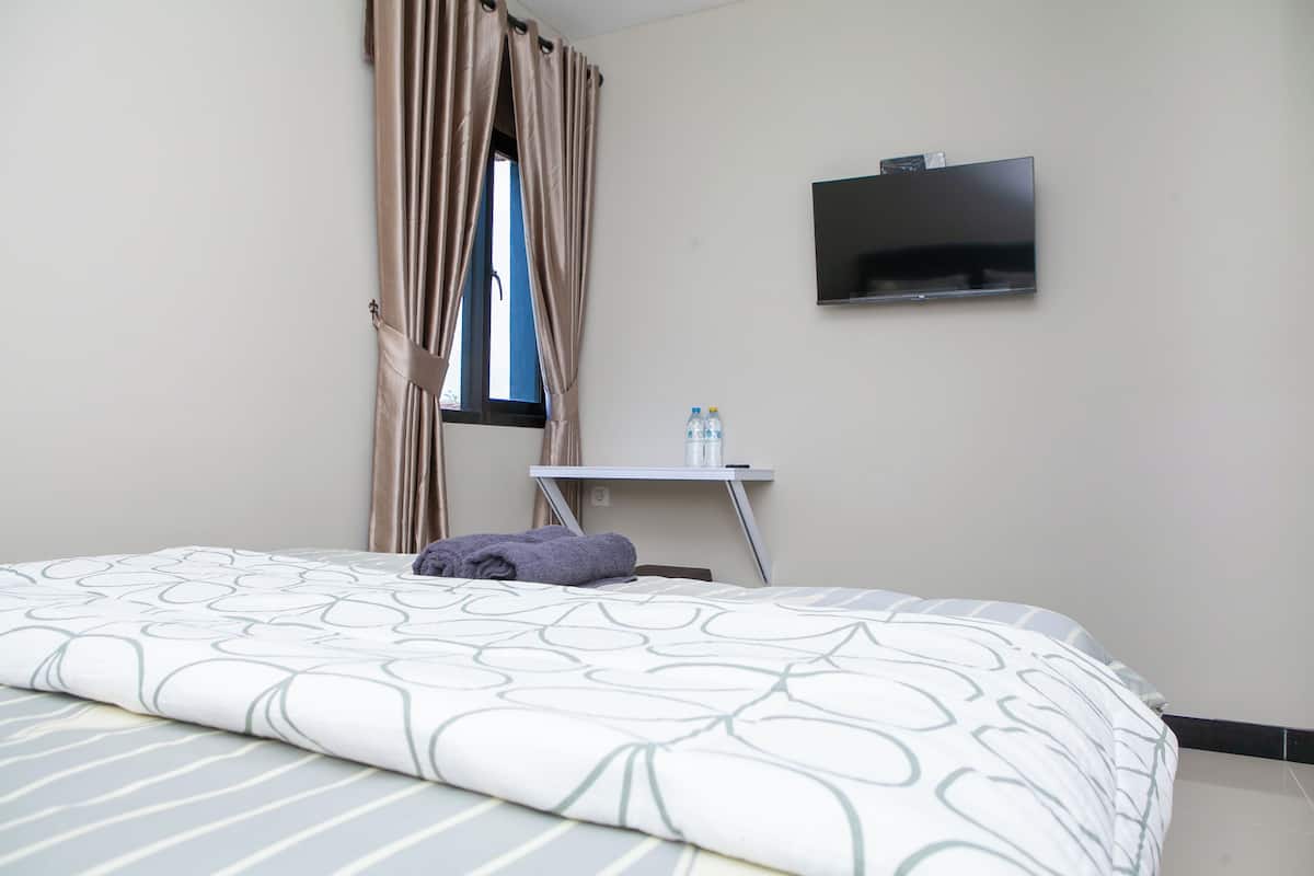 A hotel room with a bed, a TV mounted on the wall, a small table with a bottle and a glass, and a window with curtains.