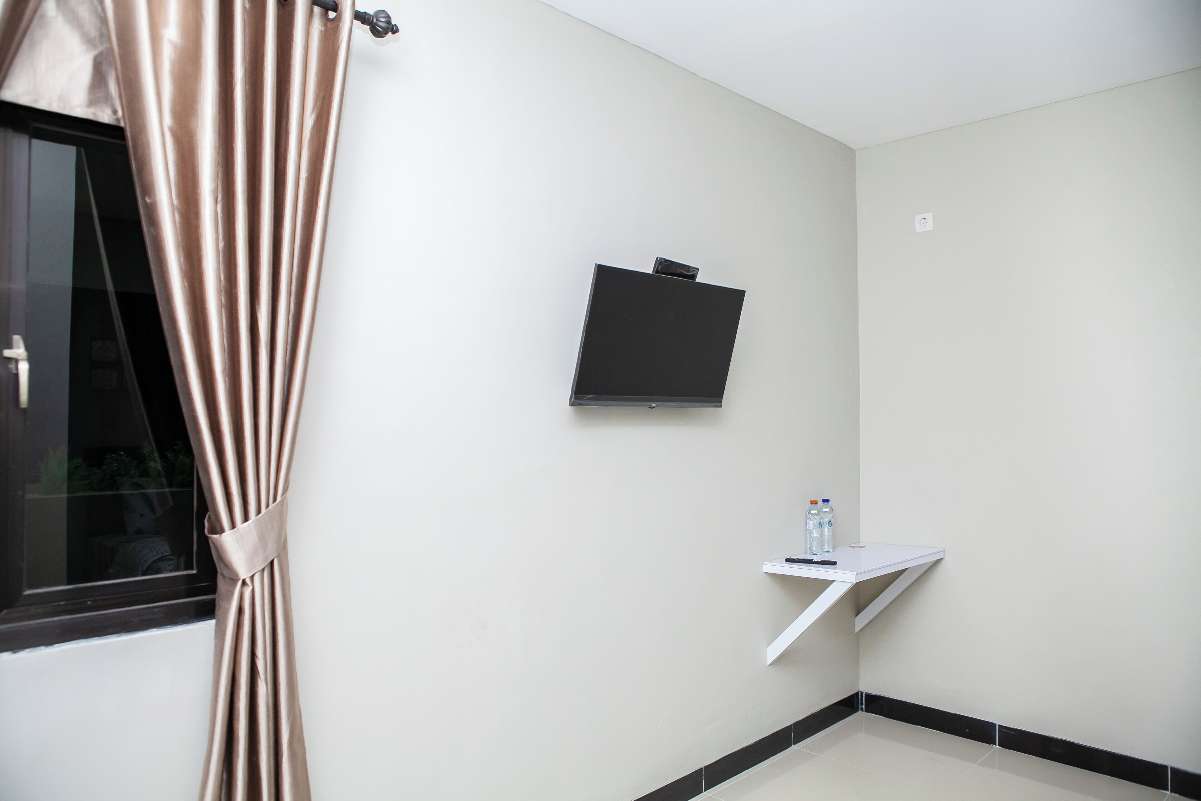 A room with a window, a mounted TV, a small white shelf with bottles, and a curtain.