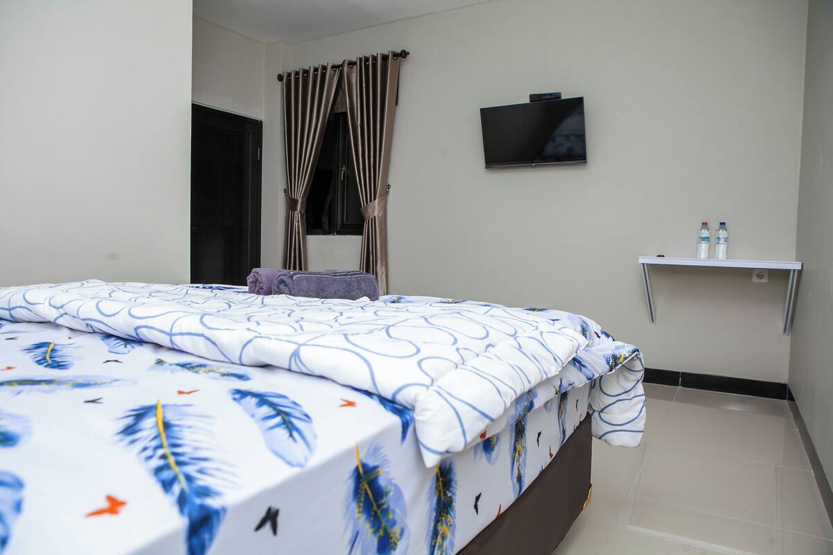 A hotel room with a bed, a television on the wall, and a small shelf with bottles.