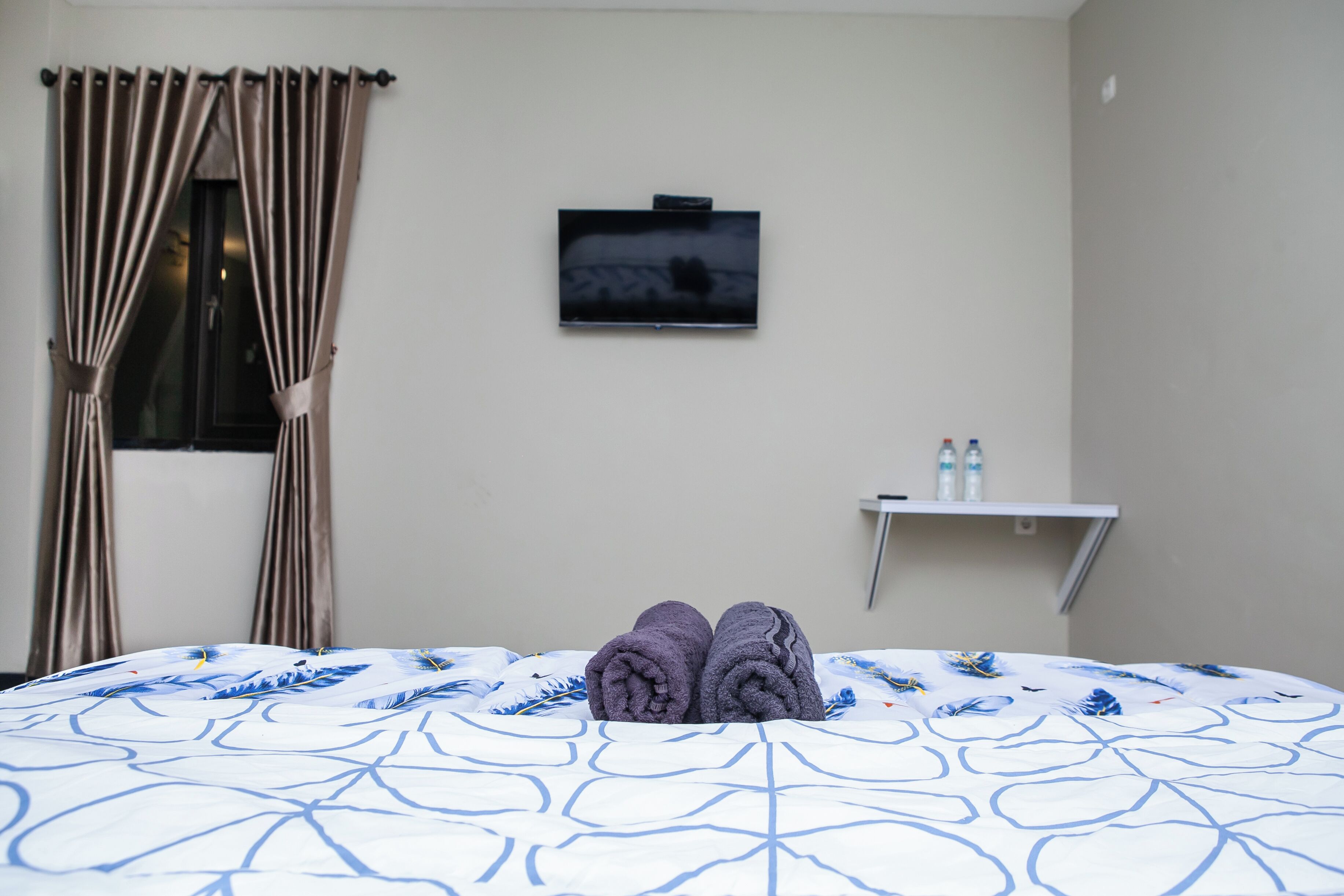 A hotel room with a bed, a TV on the wall, and a small shelf with bottles.