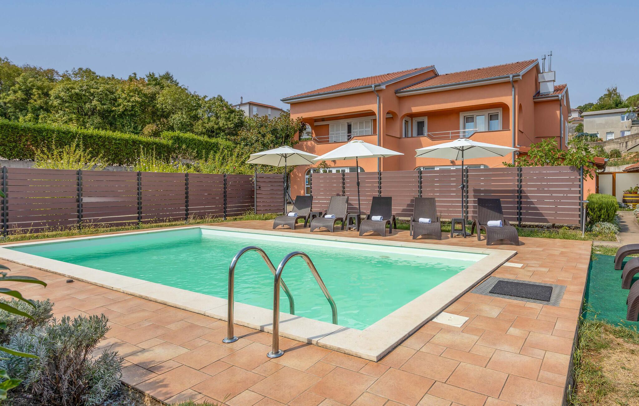 Une piscine rectangulaire avec deux échelles en métal, entourée d’une terrasse carrelée. On y trouve six chaises longues avec des parasols, une clôture en bois et une maison en arrière-plan.