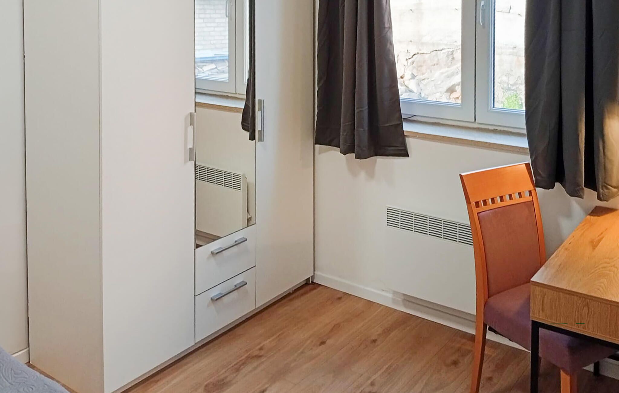 Un dormitorio con piso de madera, una cama, un armario, una silla y una ventana con cortinas.