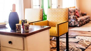 A room with a wooden desk, a vase, and two patterned mugs. There is a yellow armchair with a geometric pattern and a floral sofa in the background.
