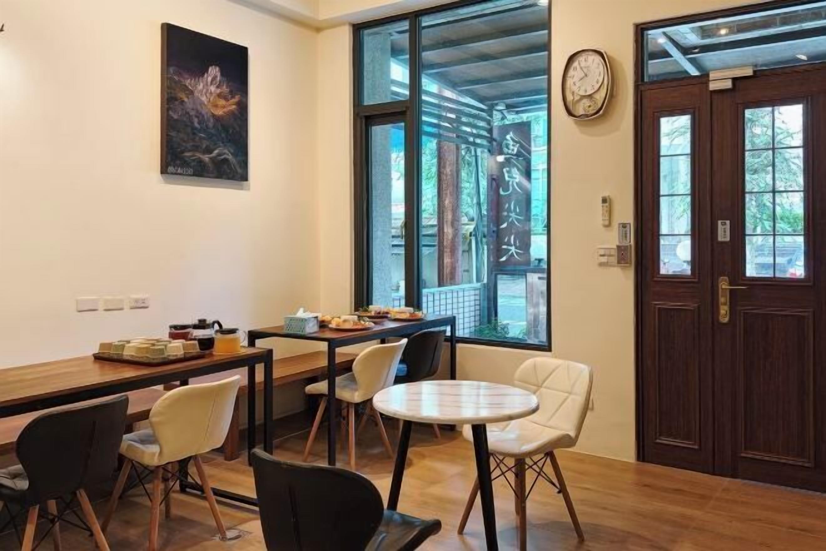 A room with a wooden floor, a round table, and chairs. There is a window with a view of trees and a door with a clock on the wall.
