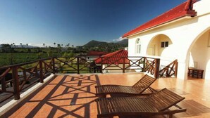 A terrace with two wooden lounge chairs, a white building with a red roof, and a view of green fields and hills. - Bali Hostel (Hualien)