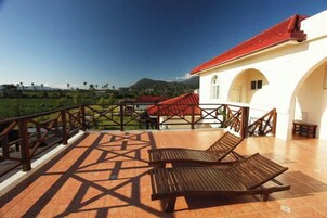 Una terraza con dos sillones de madera, un edificio blanco de techo rojo y vistas a campos verdes y colinas.