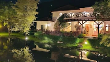 Una casa moderna con un estanque, un puente cubierto y jardines bien cuidados, todo iluminado por la noche.