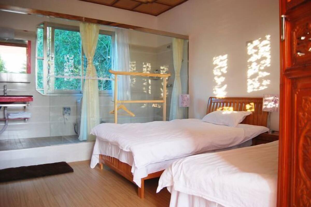 A room with two beds, a wooden headboard, a window with curtains, and a view of greenery outside.