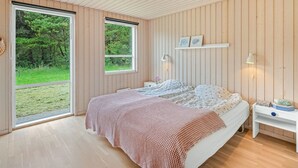 Ett sovrum med ett stort fönster, träväggar, en säng med ett mönstrat överkast, ett sängbord med en lampa och en hylla med bilder.