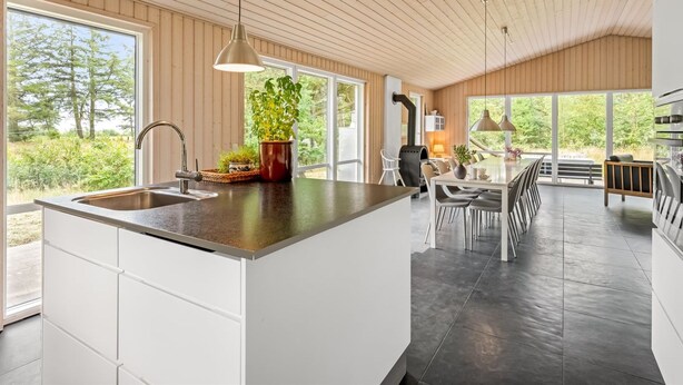 Ett modernt kök med en stor köksö, träpanel i taket och stora fönster som ger utsikt över omgivningen.