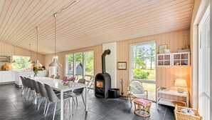 Ett modernt kök med en central köksö, en matplats med stolar, en öppen spis och stora fönster med utsikt över omgivningen.