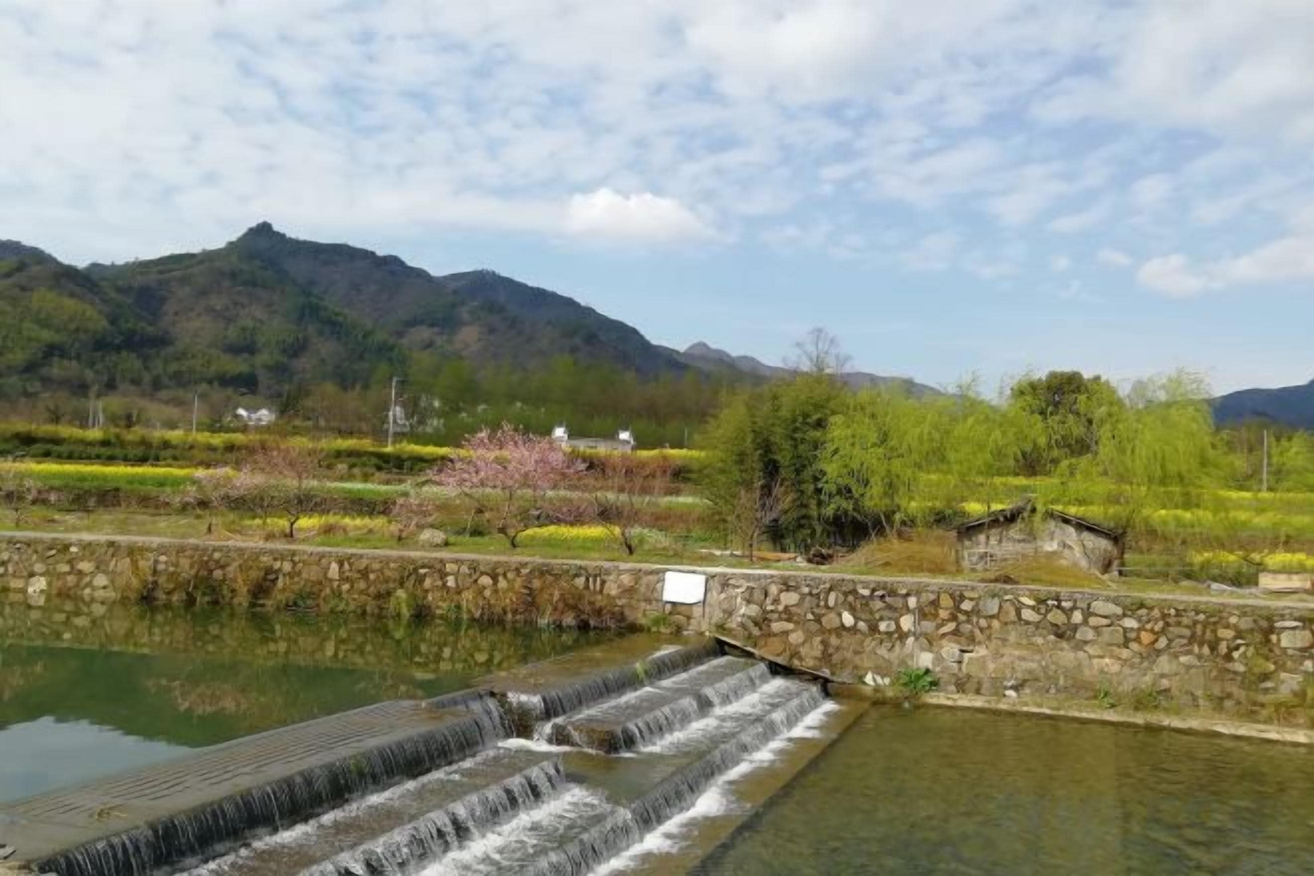 A stone bridge over a calm river with a waterfall, surrounded by lush greenery and mountains in the background.