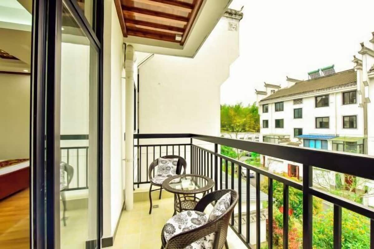 Un balcon avec des fauteuils en osier et une table en verre, donnant sur un jardin et les bâtiments voisins.