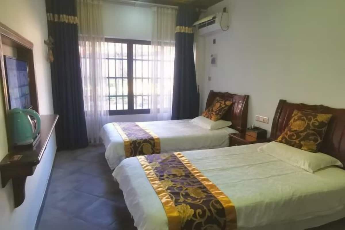 Une chambre d’hôtel avec deux lits, une fenêtre avec des rideaux, un climatiseur fixé au mur et une petite table sur laquelle se trouve un objet vert.