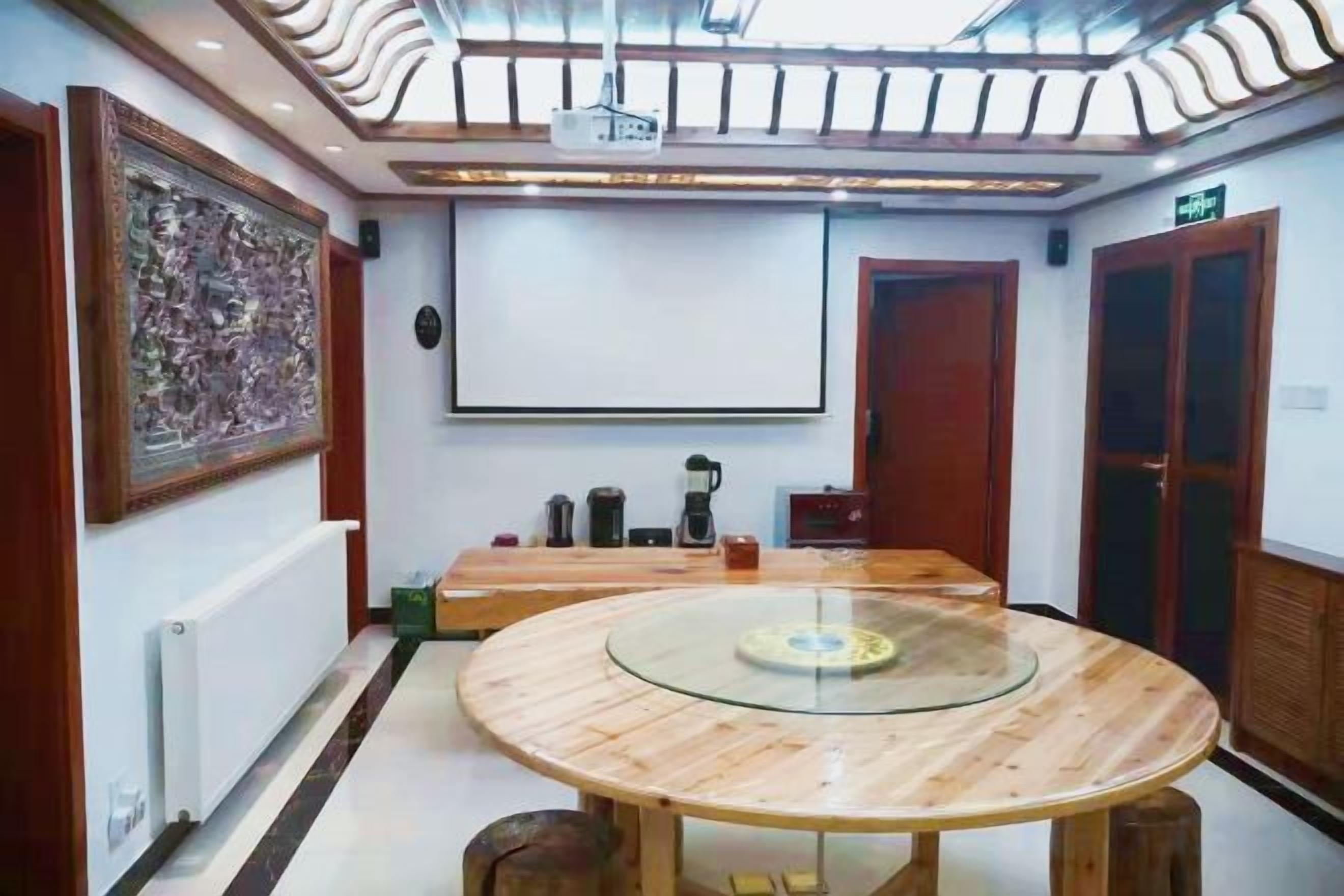 A conference room with a round glass table, wooden chairs, a projector screen, and a large painting on the wall.