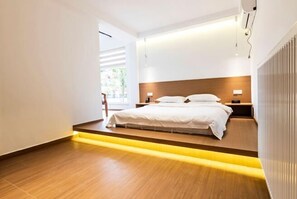 Una habitación de hotel moderna con piso de madera, una cama con ropa blanca, un mesita de noche y una lámpara.