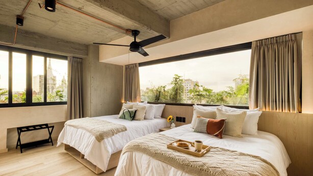 Habitación de hotel con dos camas, ventilador de techo, ventanas amplias y vistas a zonas verdes y edificios.