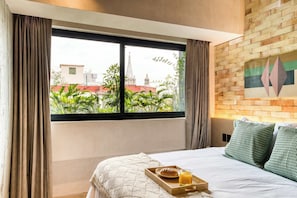 Une chambre d’hôtel avec un lit, une fenêtre avec des rideaux, un plateau avec une pâtisserie et du jus de fruits, et un mur en briques orné d’un tableau aux motifs géométriques.