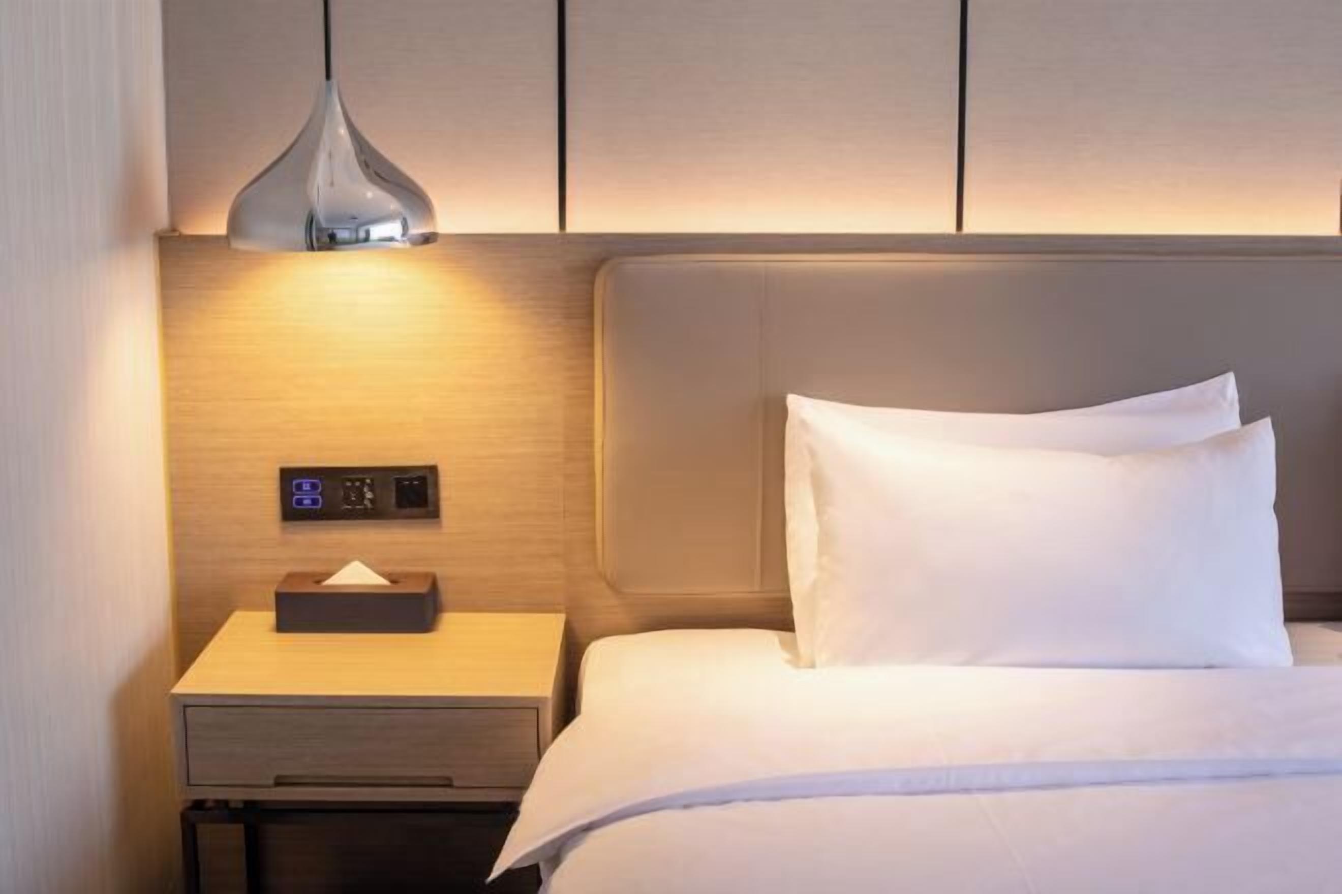 A hotel room with a bed, a bedside table with a lamp, a control panel, and a drawer.