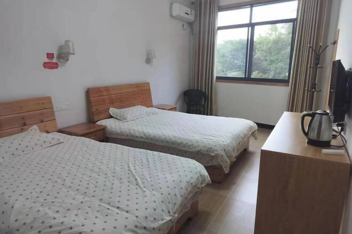 Quarto de hotel com duas camas, uma mesa de cabeceira de madeira, uma escrivaninha com chaleira, televisão e janela com cortinas.