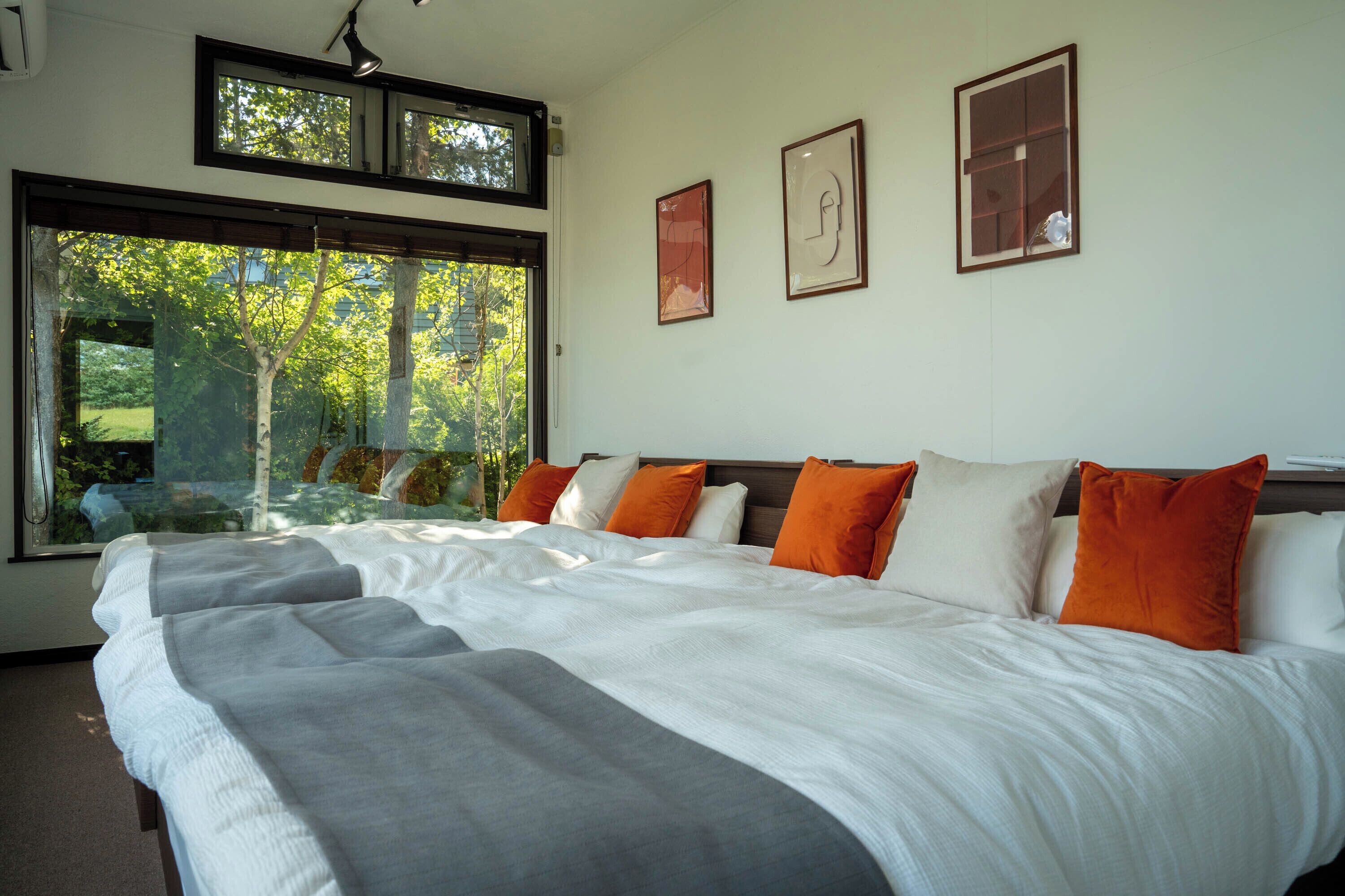 Un grand lit avec une literie blanche et grise, des coussins orange et blancs, et une vue sur les arbres à travers une grande fenêtre.