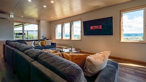 Un salon moderne doté d’un grand canapé d’angle, d’un téléviseur à écran plat diffusant du contenu de « NETFLIX », et de grandes fenêtres offrant une vue sur l’extérieur.