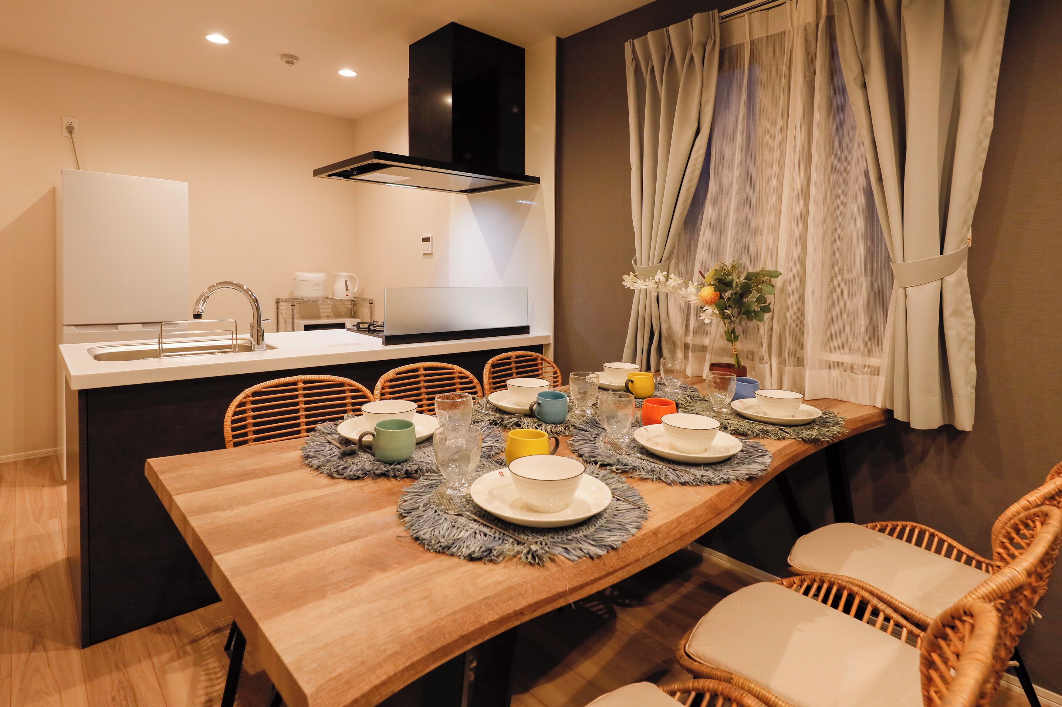 A dining area with a wooden table set for four, a kitchen in the background, and a window with curtains.