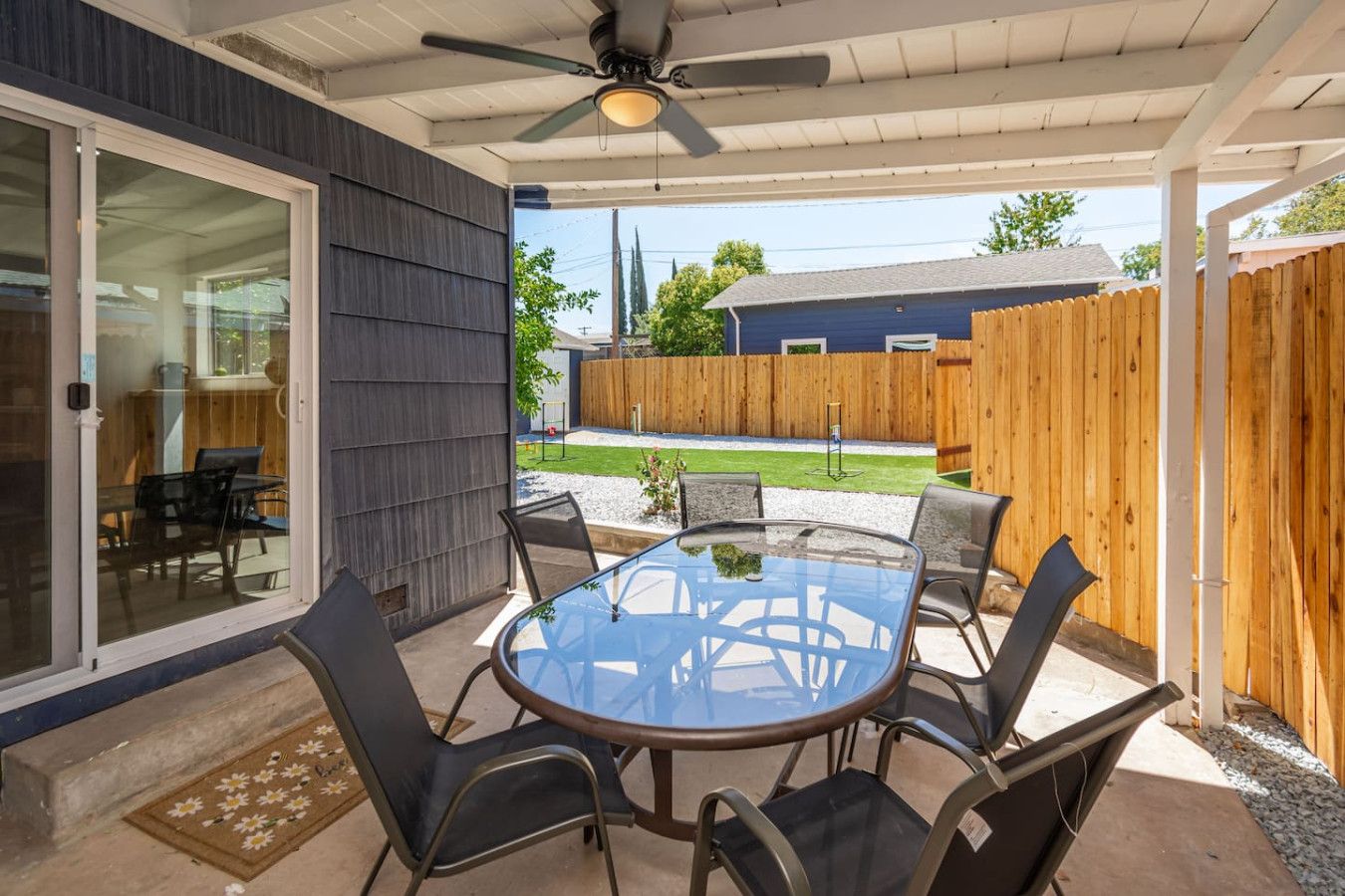 A covered patio with a glass table and chairs, a ceiling fan, and a view of a backyard with a wooden fence and a house.