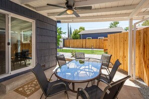 A covered patio with a glass table and chairs, a ceiling fan, and a view of a backyard with a wooden fence and a house. - Main Street Retreat in Roseville (Roseville)