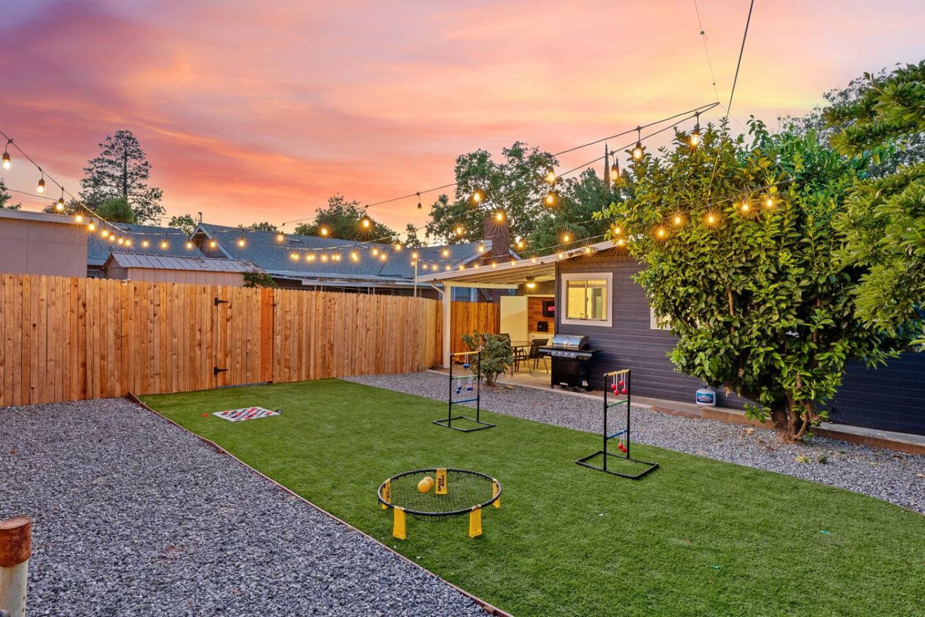 Un jardin avec une pelouse artificielle, un trampoline, un espace barbecue et une clôture en bois.