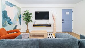 A modern living room with a grey sofa, orange armchair, and a flat-screen TV mounted on the wall. - Sojourn's Harborage - Cove Suite (Norfolk)