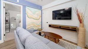 Ein Wohnzimmer mit grauem Sofa, einem runden Holztisch, einem flachbildfernseher an der Wand und einer dekorativen Vase mit Zweigen.
