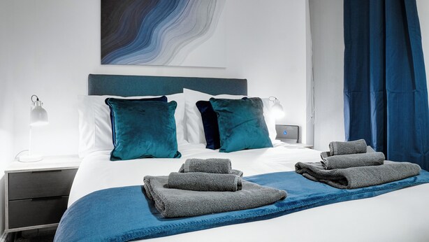 A modern hotel room with a neatly made bed, a blue throw blanket, and a large abstract painting on the wall.