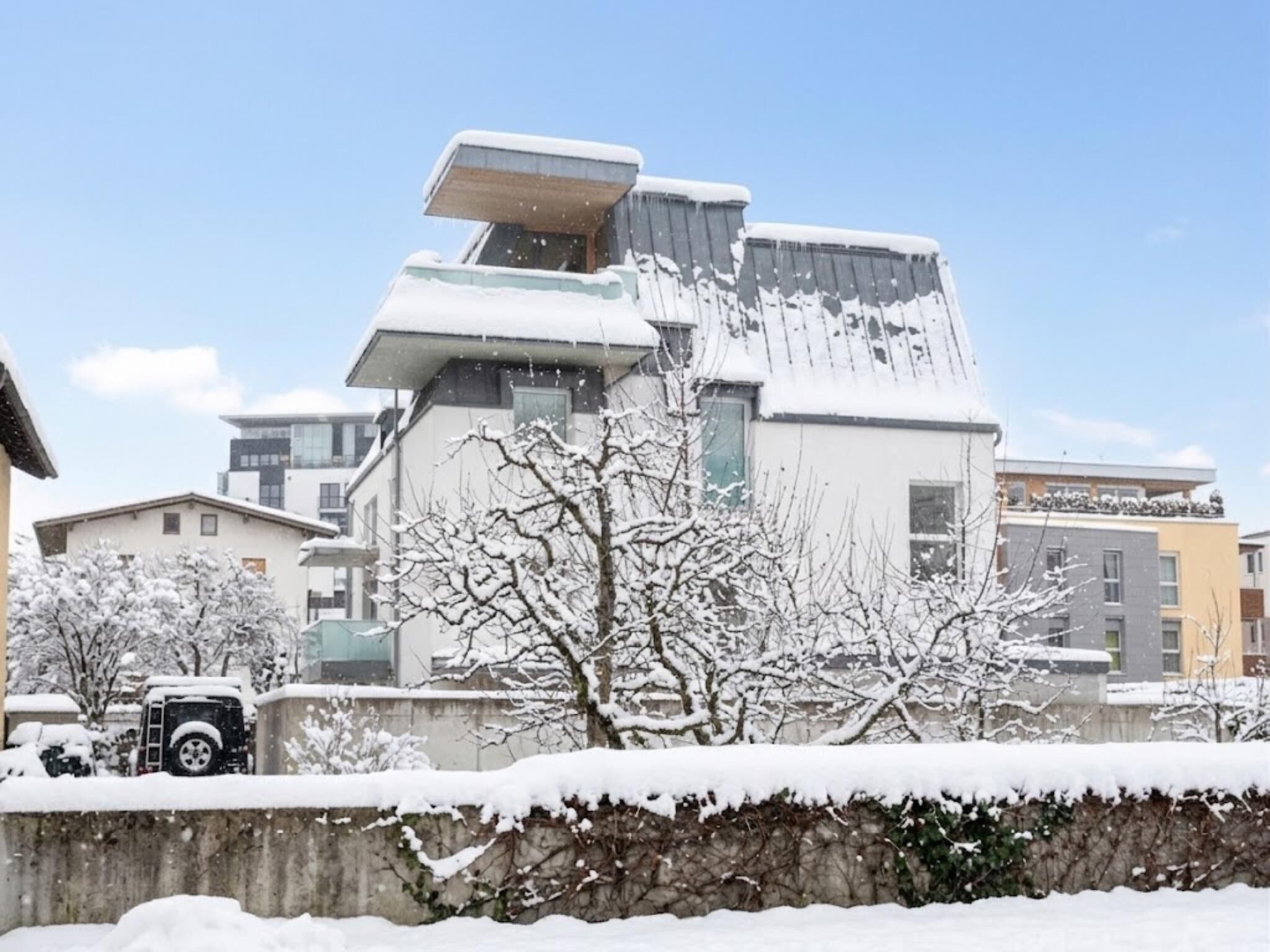 Un edificio moderno con techo plano y amplios ventanales, rodeado de árboles cubiertos de nieve y un muro de piedra.
