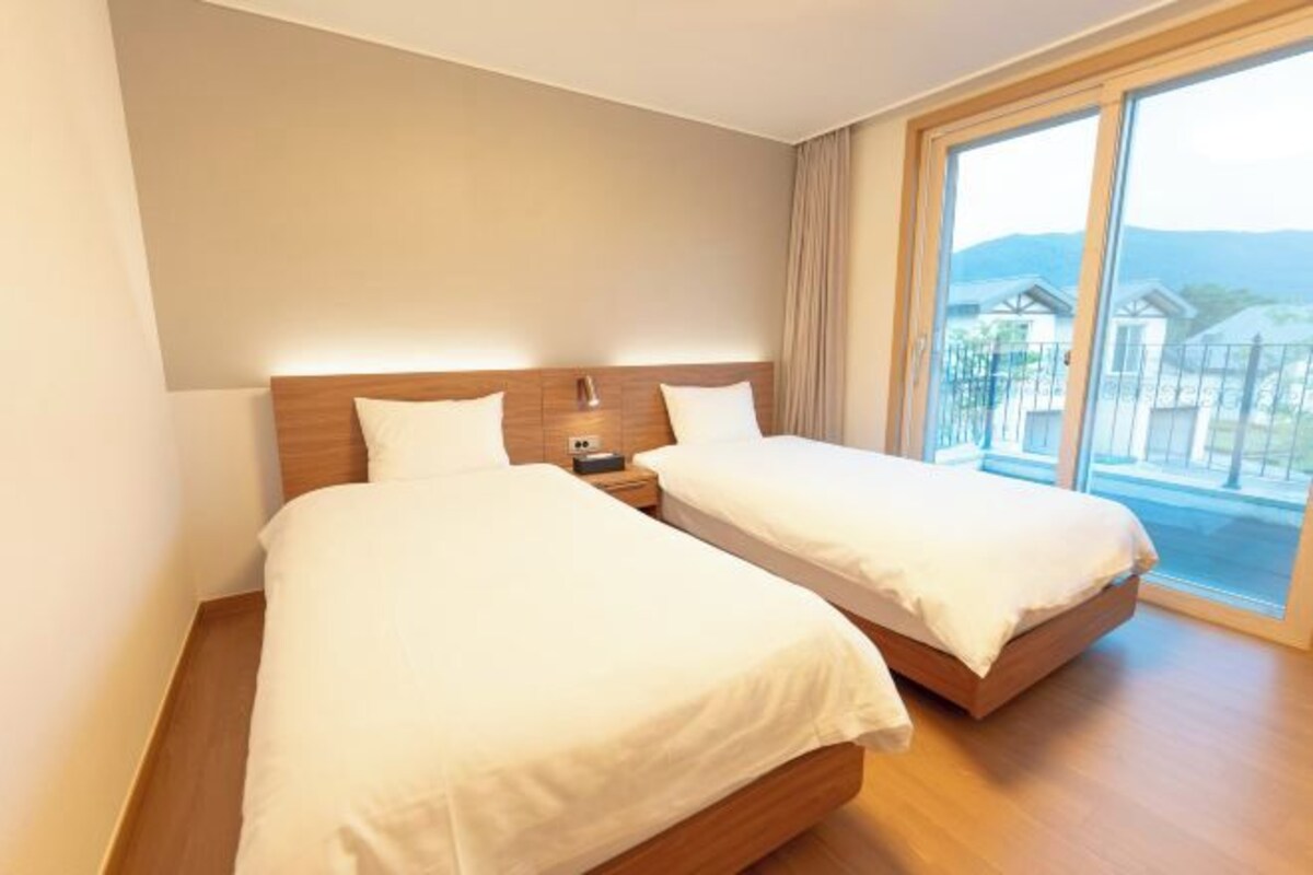 Ein Hotelzimmer mit zwei Betten, einem Holzkopfteil, einem Nachttisch und einem großen Fenster mit Blick auf Gebäude und Berge.