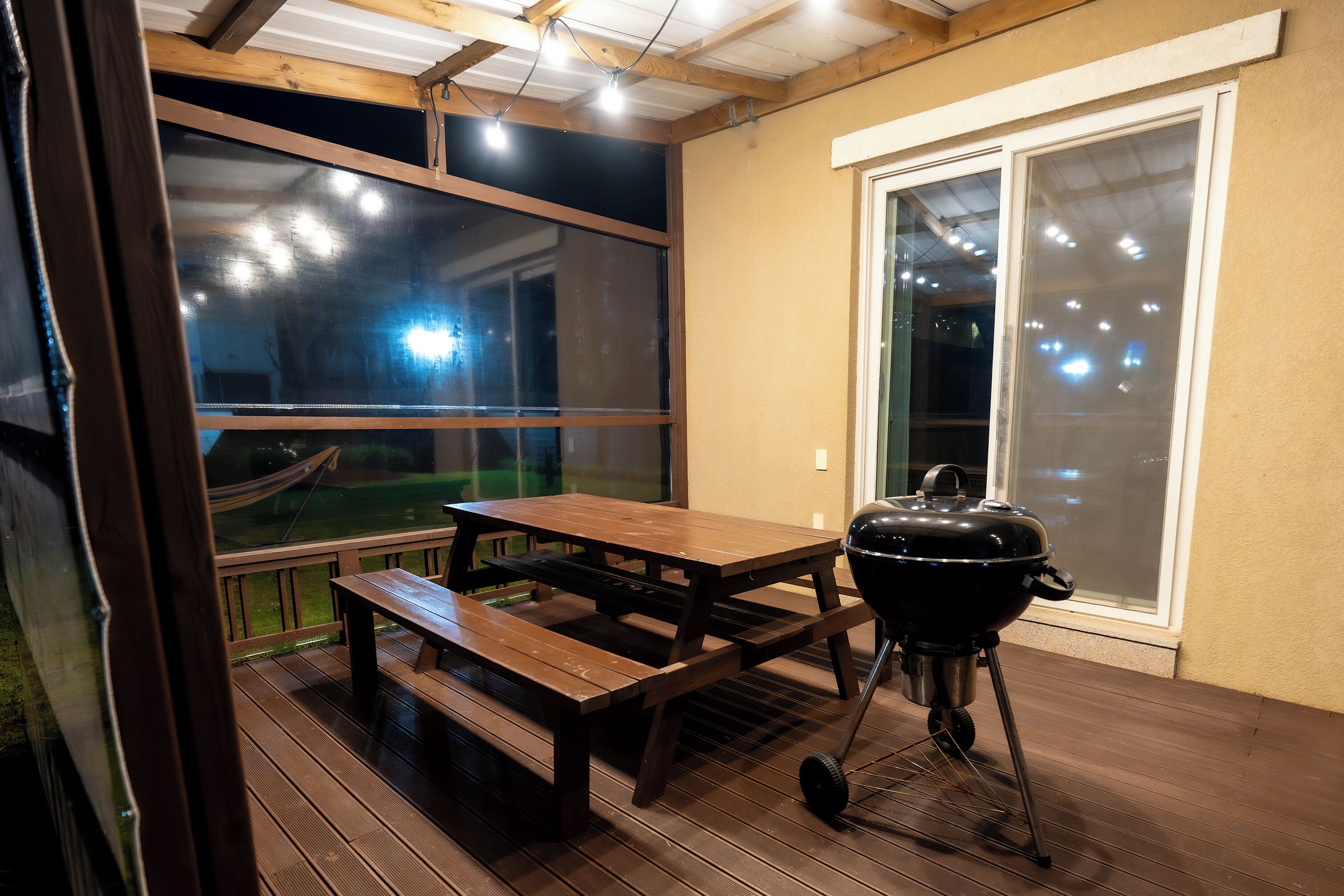 Un patio exterior con una mesa y bancos de madera, una parrilla y vistas a través de una ventana.