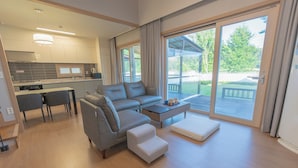 Un salon moderne avec un canapé gris, un pouf blanc et une table basse. Il y a également un coin cuisine avec une table à manger et des chaises. De grandes fenêtres offrent une vue sur l’extérieur.