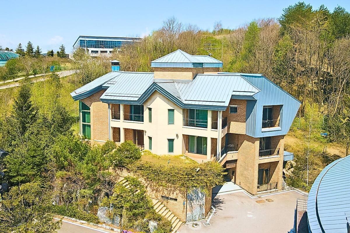 A modern multi-story building with a flat roof, surrounded by trees and a clear sky.