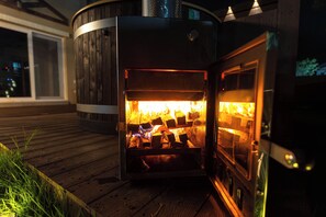 A wood-burning stove with visible logs inside, set on a wooden deck. - Le Comte Bleu (Pyeongchang)