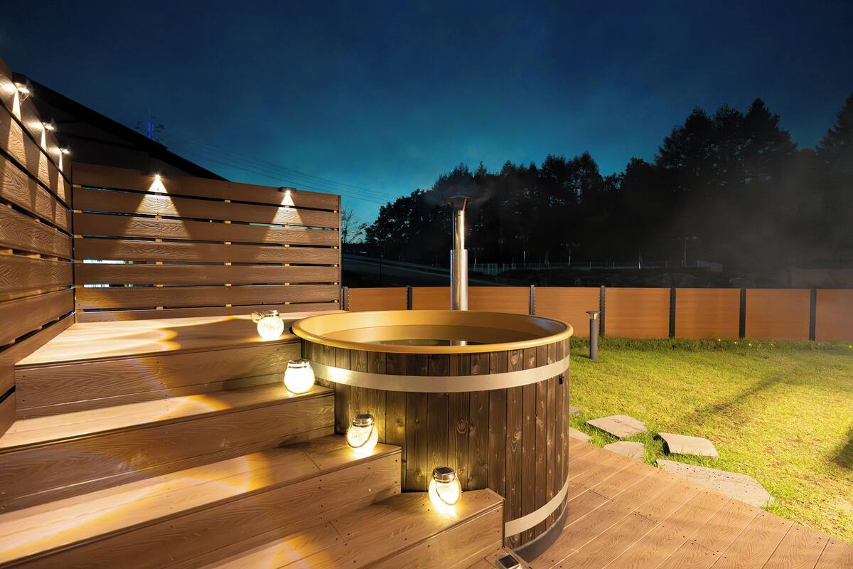 A hot tub with wooden steps and lighting, surrounded by a wooden deck and a grassy area.