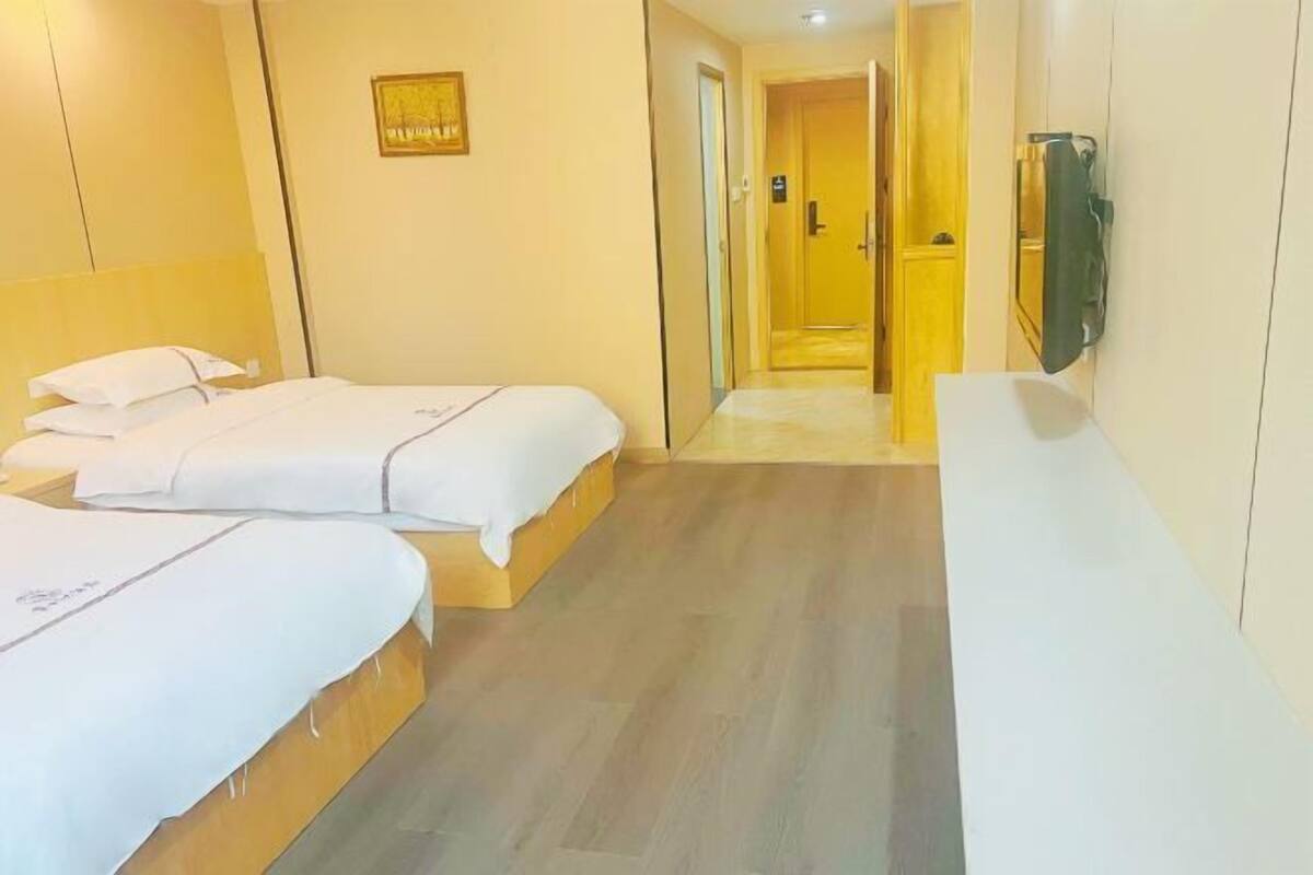 A hotel room with two beds, a wooden headboard, a flat-screen TV mounted on the wall, and a hallway leading to another room.