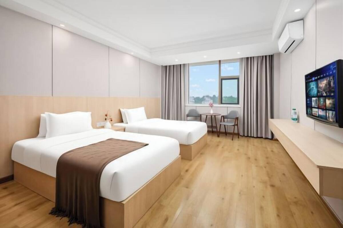 A hotel room with two beds, a small table, a chair, a TV, and a window with curtains.