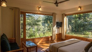 A bedroom with a bed, a desk, and a chair. There are large windows with curtains, a ceiling fan, and a wall-mounted TV.